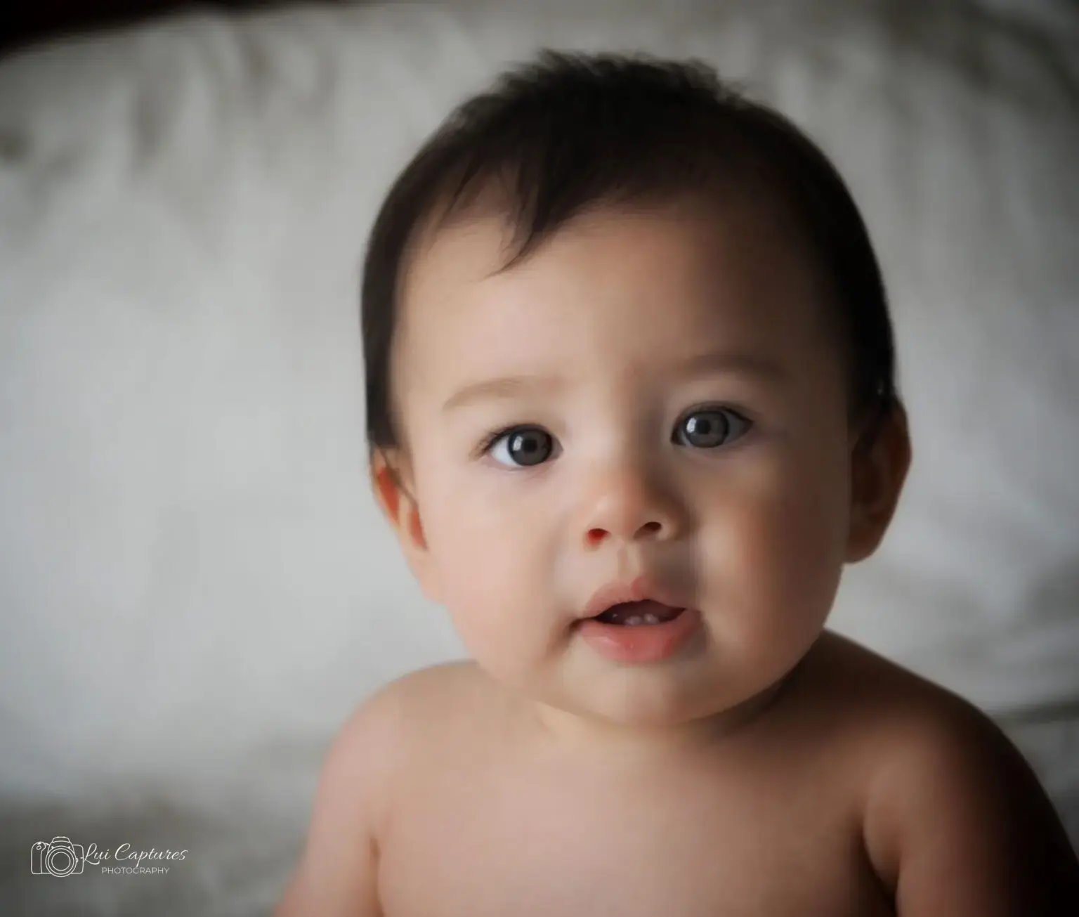 Close-up portrait of a baby with light brown skin and dark brown hair. The baby's big, round eyes are looking directly at the camera, showing a curious and gentle expression. The baby is shirtless, highlighting their soft, baby skin against a blurred light grey background. A subtle smile is visible, enhancing the baby's innocent and adorable appearance.