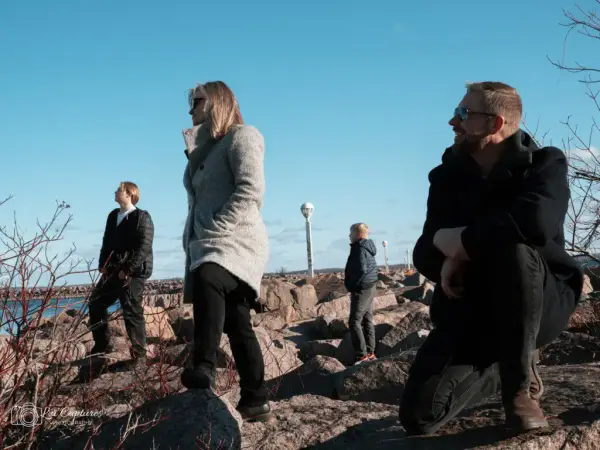 Alt text: "A family of four scattered on a rocky lakeshore under a clear blue sky. From left to right, a young boy and a woman are standing, each gazing out over the lake, while a younger boy explores nearby. On the right, a bearded man in a black coat crouches on the rocks, looking contemplatively at the water. The scene captures a moment of peaceful individual reflection amidst the rugged natural beauty of the Kingston, Ontario lakeside environment.