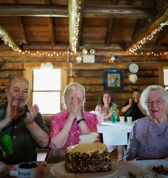 A heartwarming moment captured in birthday and event photography, showing an elderly woman in a pink blouse, visibly moved and covering her face with her hands in joy at a birthday celebration. Beside her, a man in green and another elderly woman in gray applaud with smiles. The scene is set in a cozy, rustic log cabin adorned with string lights, adding a festive atmosphere. In front of them is a large birthday cake on the table, making the moment even more special.